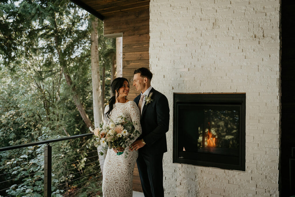 The bride and groom smiling at each other at their Airbnb by the fireplace. 