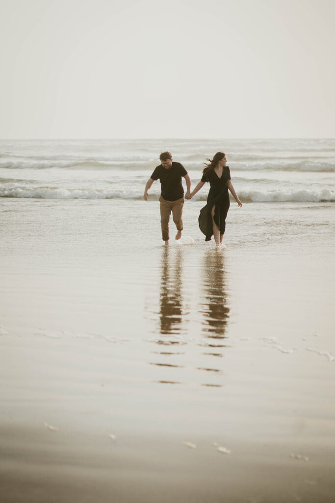 Couple holding hands, running across Cannon Beach during their beach engagement photos