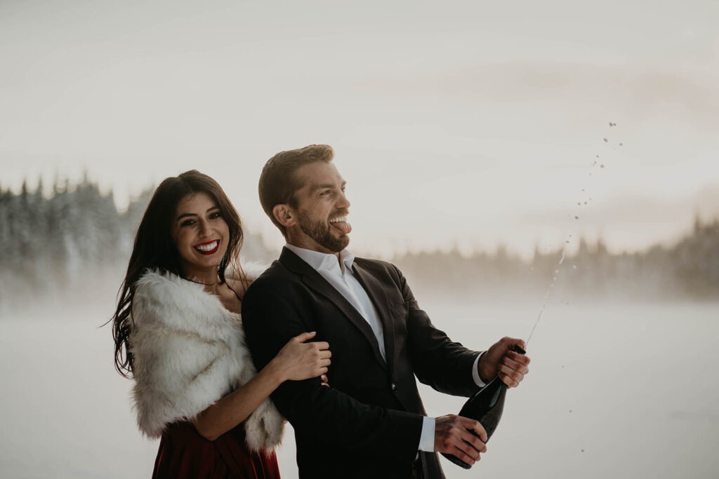 Couple popping champagne during engagement photos in the snow