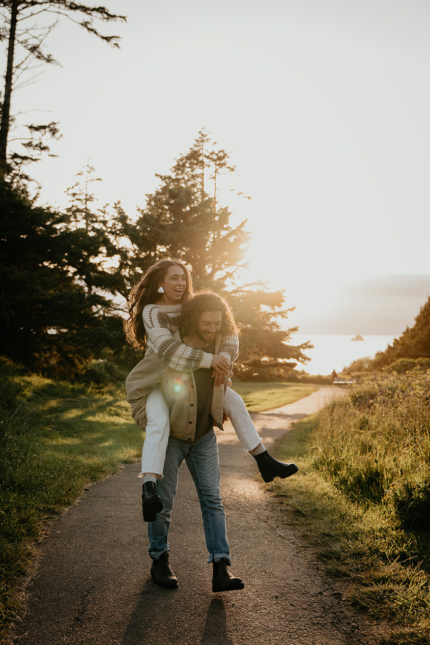 a boyfriend carrying his girlfriend on his back hiking along a coastal path during their engagement photo session.