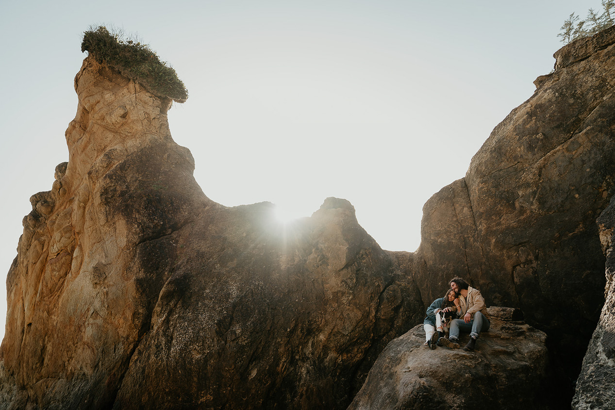 a couple and their dog hugging on a boulder along the Oregon coast during their engagement photo session. 