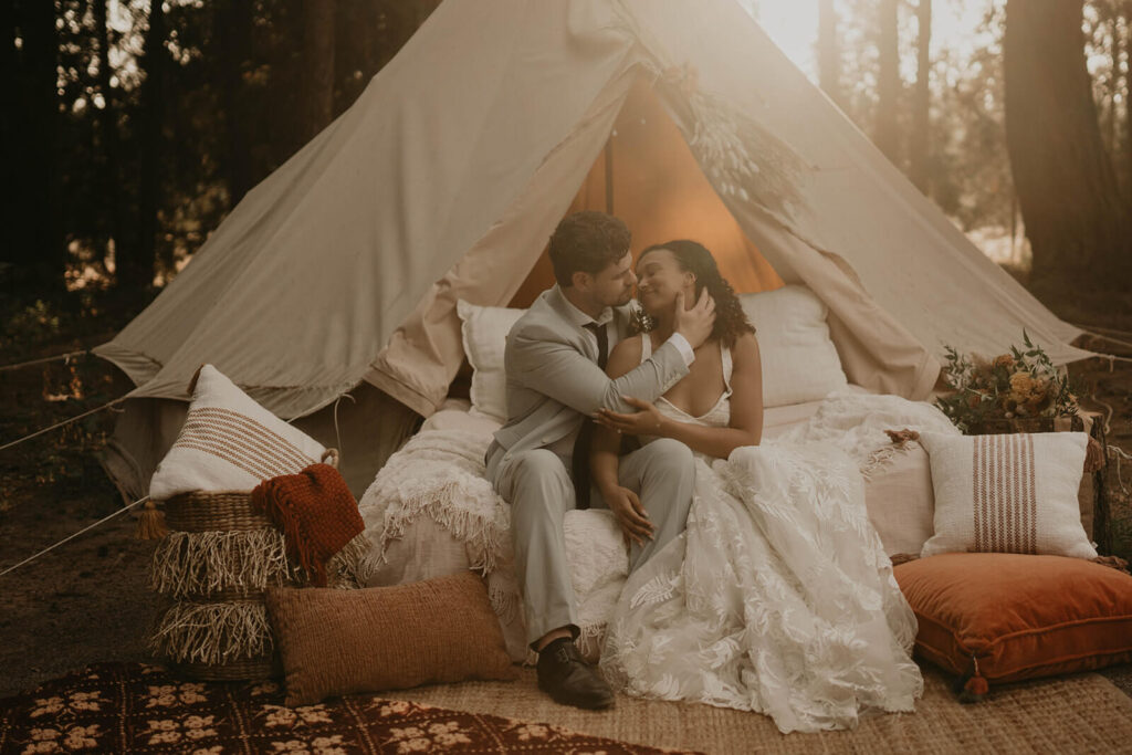 Bride and groom sitting on a stack of pillows in front of a woodland themed wedding tent