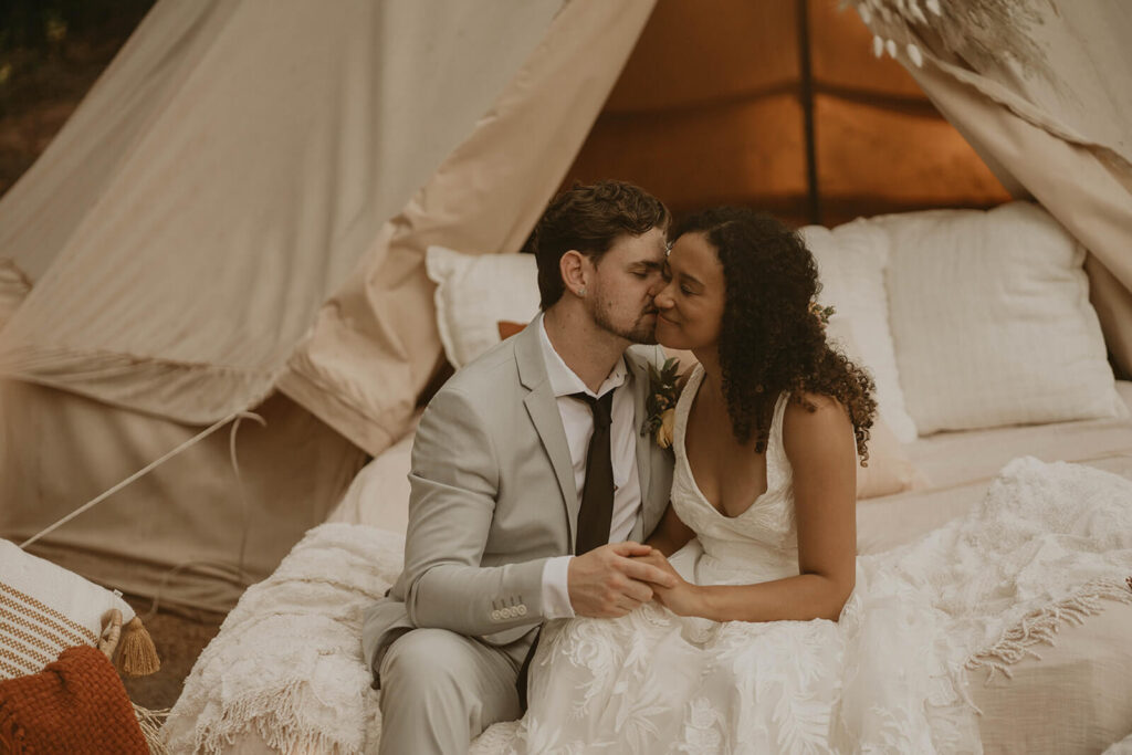 Bride and groom kiss while sitting on an outdoor bed in the woods surrounded by orange pillows and a white linen tent