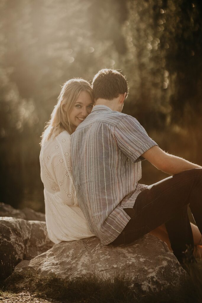 Couple sitting on a rock by a lake and woman looking over her shoulder, smiling at the camera