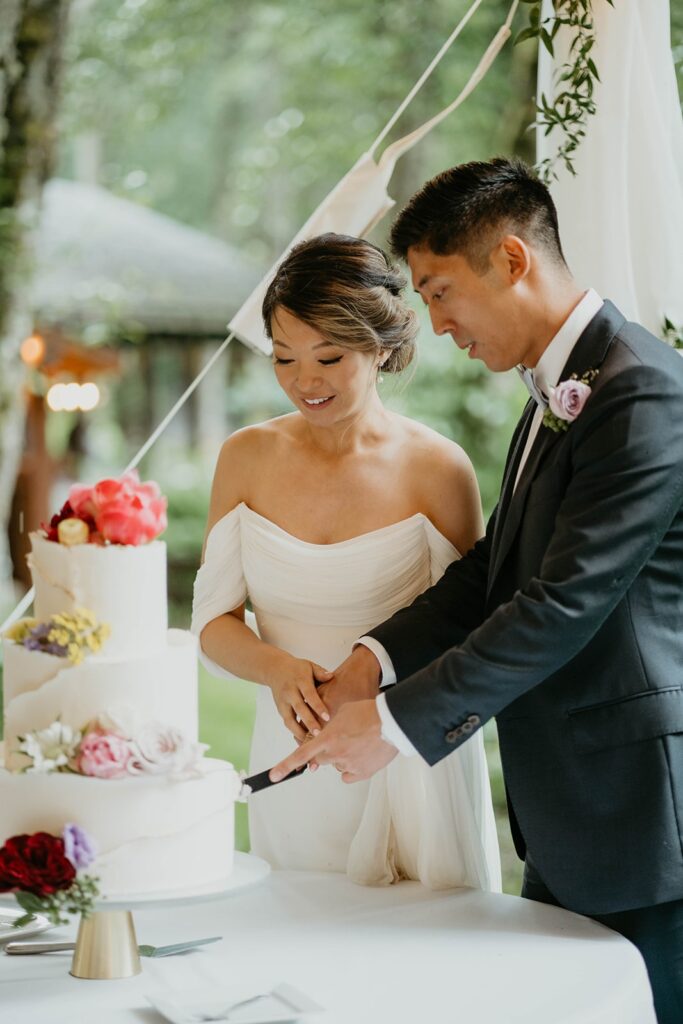 Bride and groom cut into three tier white wedding cake with flowers