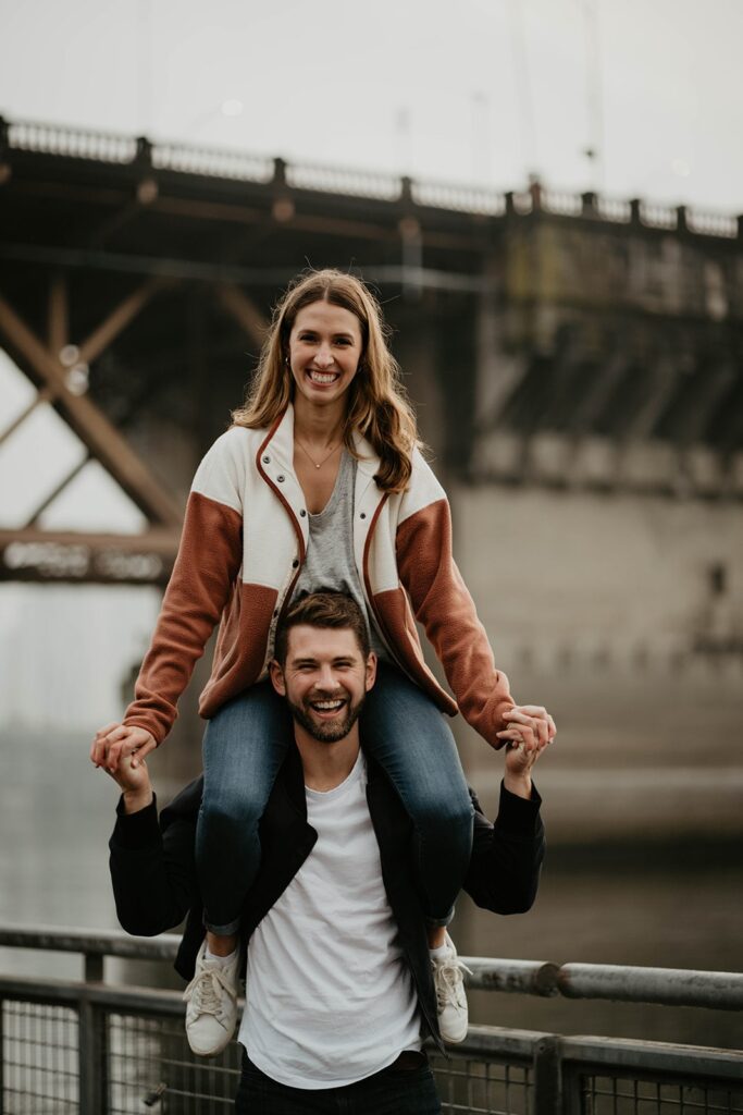 Woman riding on man's shoulders during Portland engagement photo session by the river