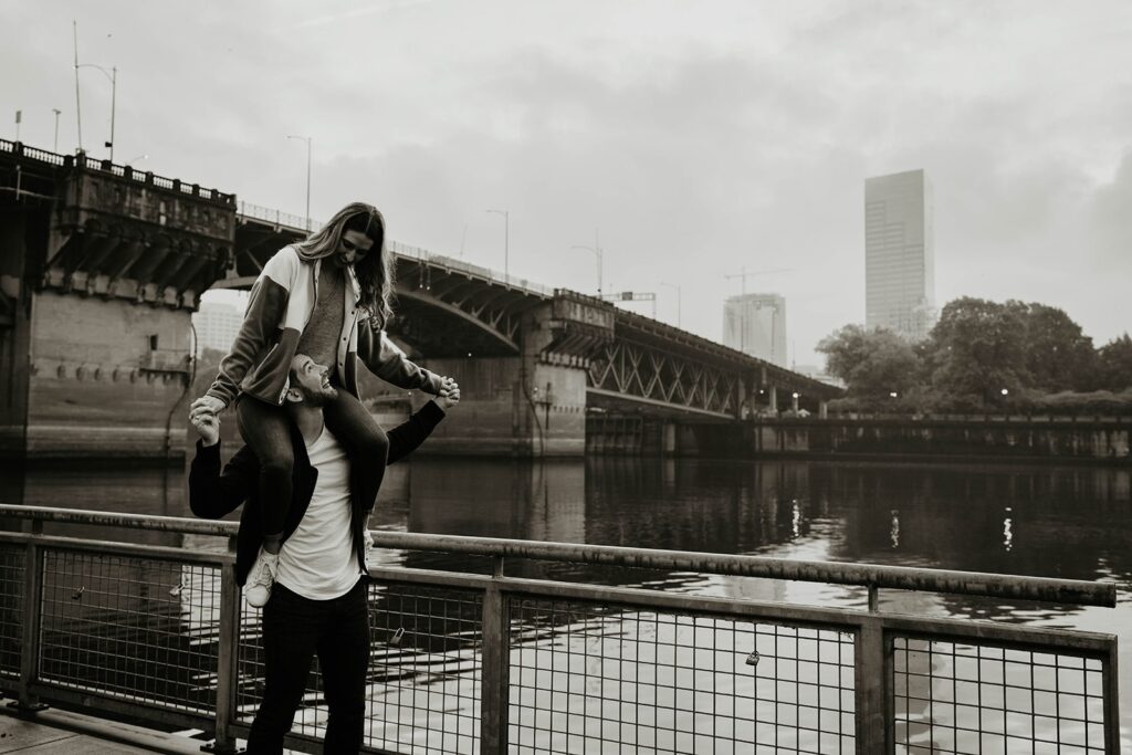 Woman riding on man's shoulders during Portland engagement photo session by the river