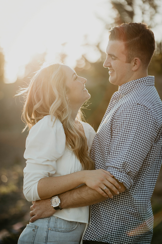 A couple smiling lovingly at each other with trees and the sun in the background at the Timberline Lodge. 