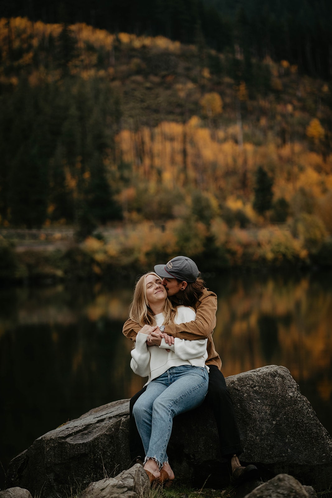 Couple sits on a rock at Government Cove while man kisses woman on the cheek at their Oregon engagement photo session