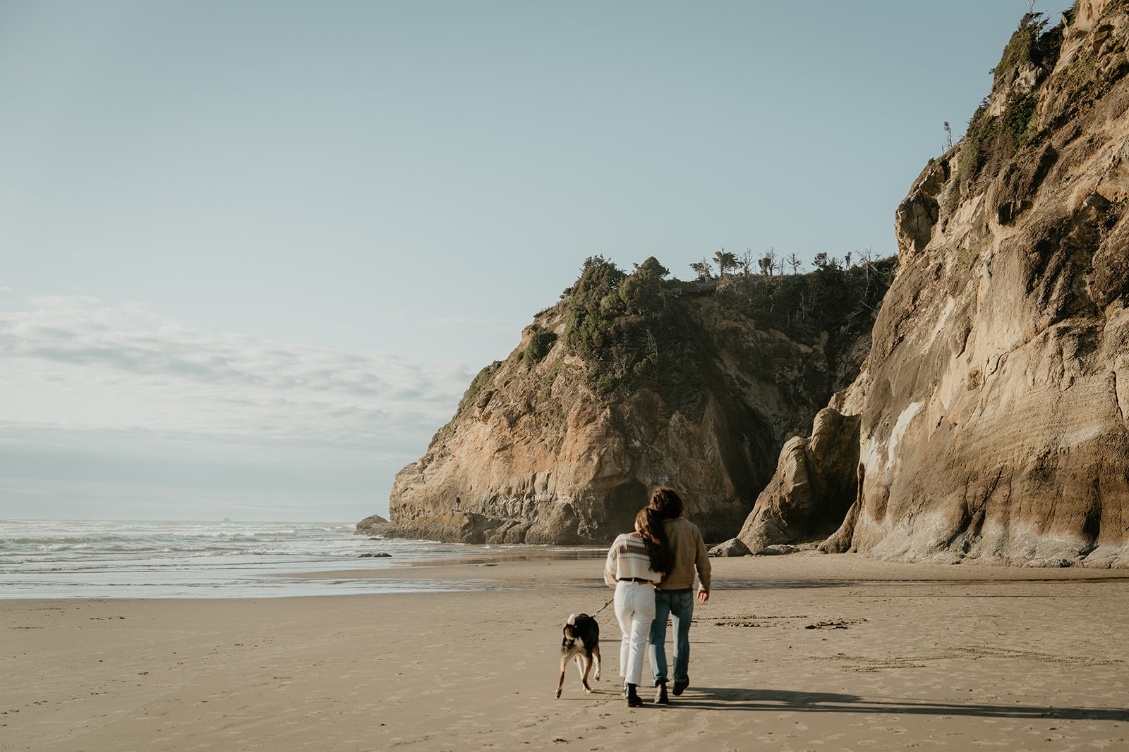 Couple links arms and walks across the beach at Hug Point during their Oregon engagement photo session