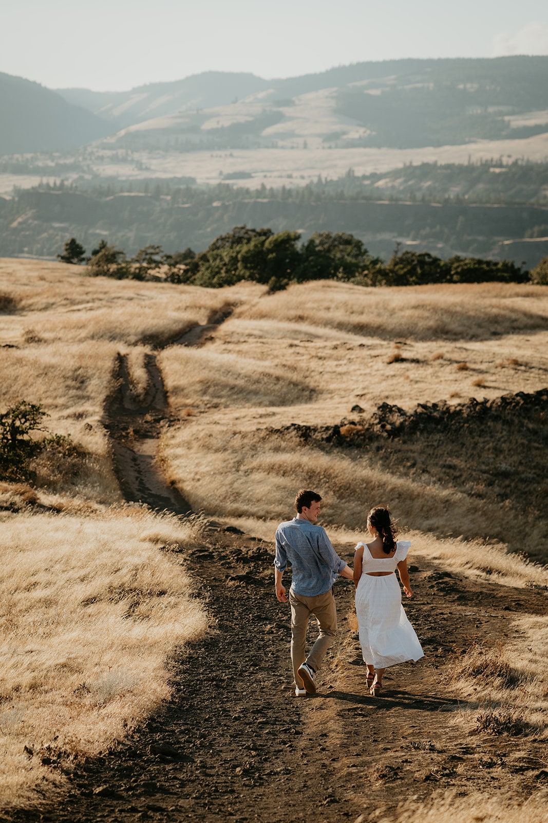 Couple holds hands while walking down a dirt path during their Oregon engagement photo session at Rowena Crest