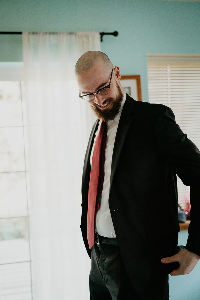 A groom wearing a black jacket, black pants, white shirt, and red tie adjusting his jacket inside a Portland Airbnb. 