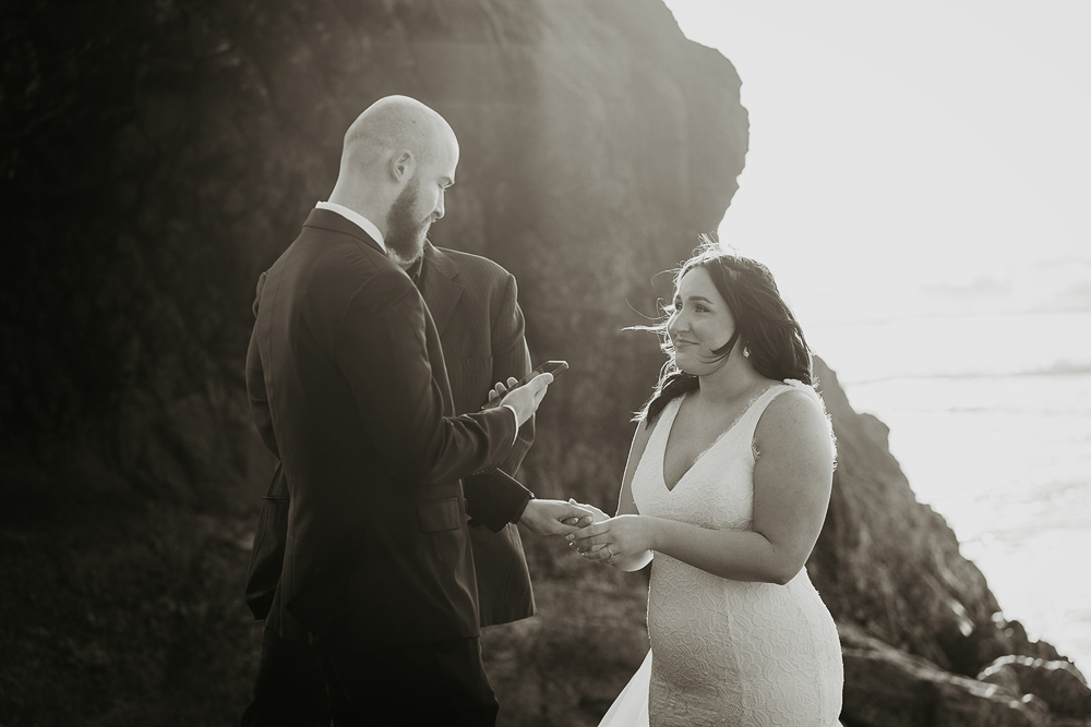 The groom reading his vows to the bride with the officiant in the background. All three are standing on a sea stack at Cannon Beach. 