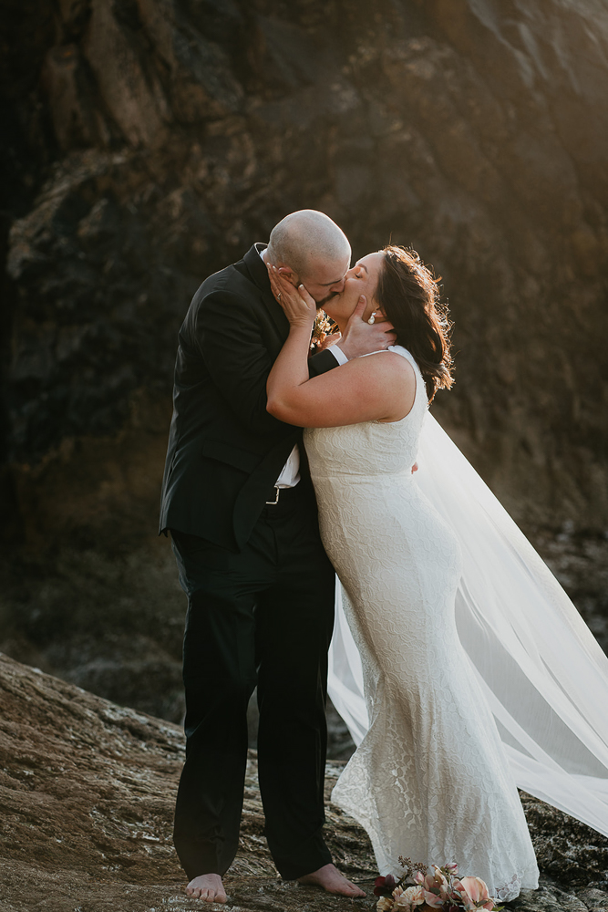 The bride and groom kissing while standing barefoot on a sea stack at Cannon Beach. 