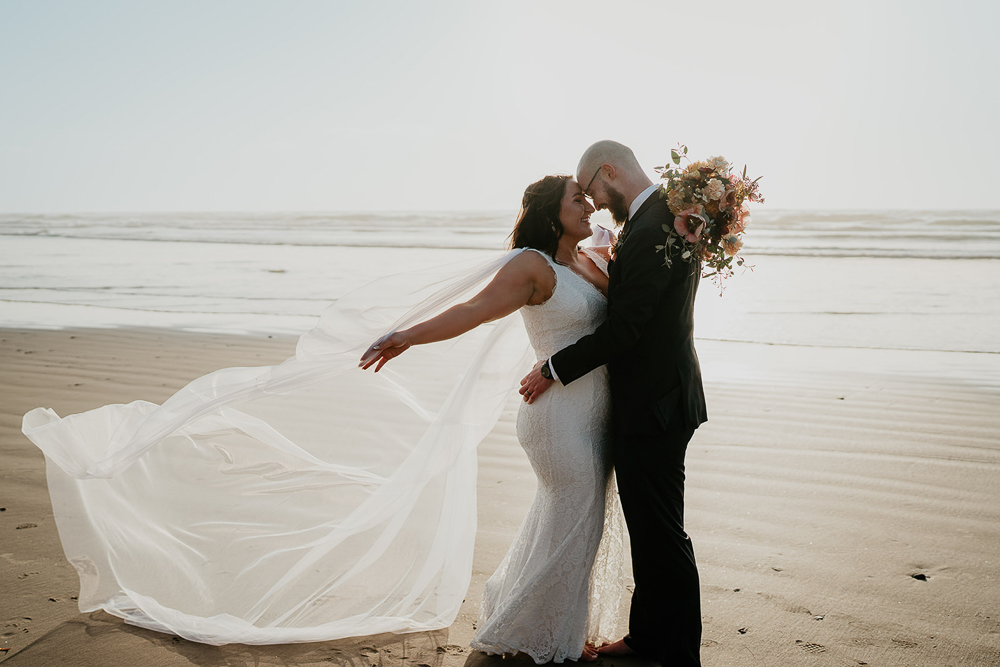 The groom holding the bride while her veil flows in the wind, with both standing on the sand at Cannon Beach with the ocean in the background. 