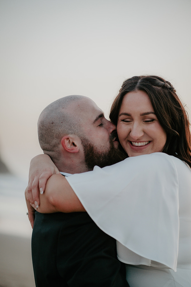 The groom giving his bride a kiss on the cheek at Cannon Beach. 