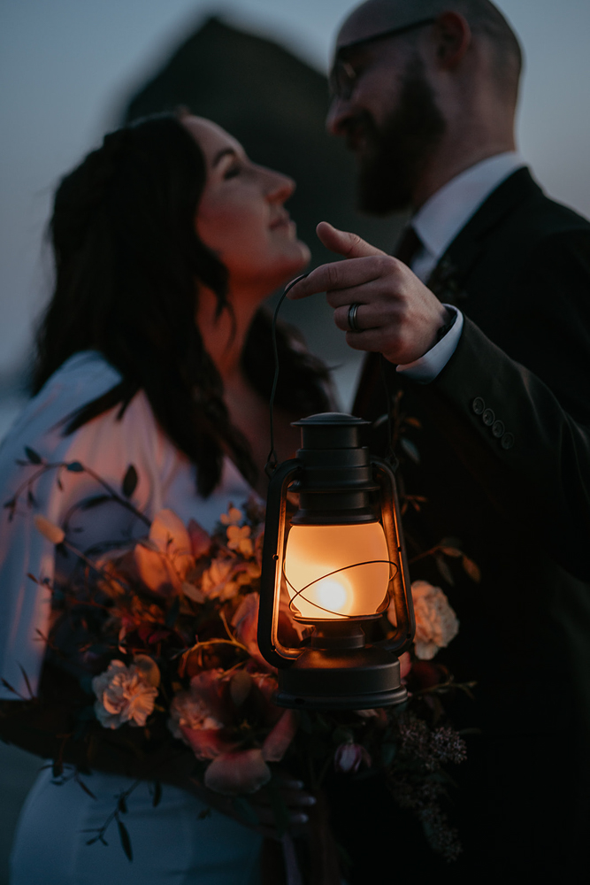 Newlyweds lit up by the a lantern and holding a flower bouquet with Haystack Rock in the background at Cannon Beach. 