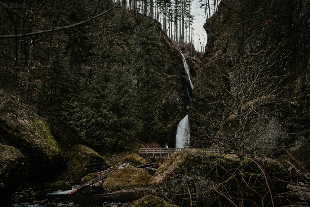 A couple standing on a bridge with mossy boulders and a river in the foreground, and a waterfall in the background at the Columbia River Gorge. 