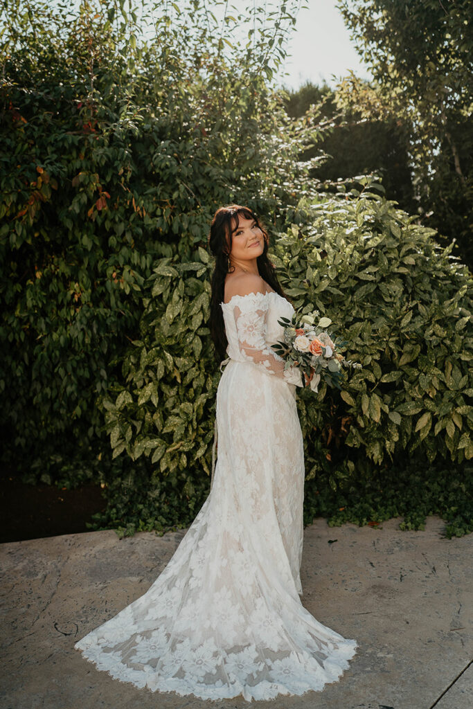 The bride standing among greenery and holding a bouquet. 