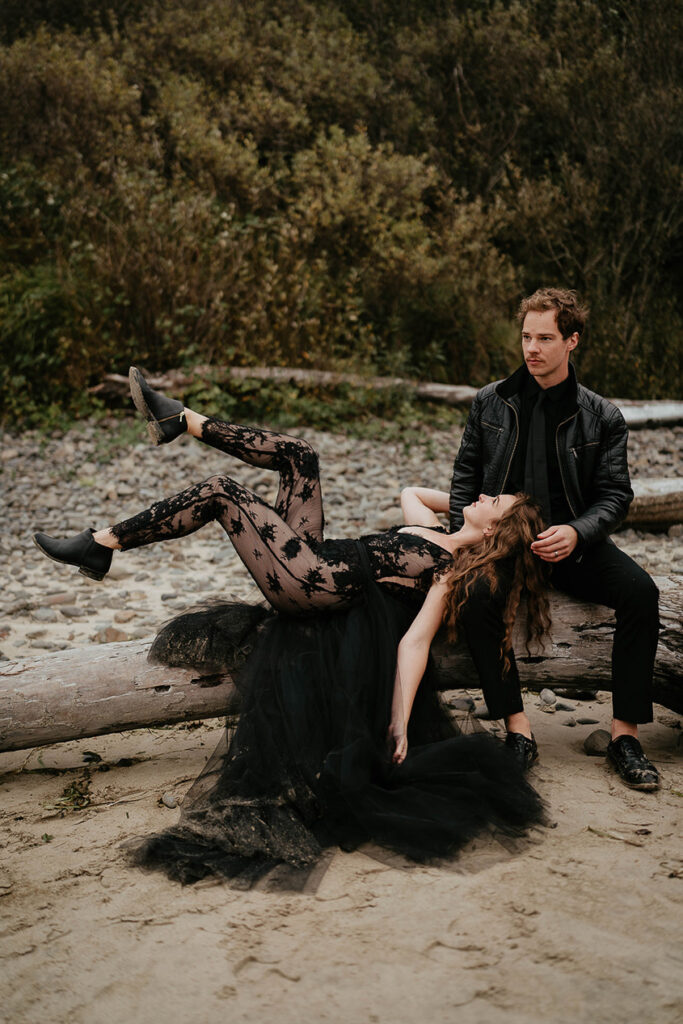 A man sitting on a log, with his partner's head resting on his leg while she lays down on the log. 