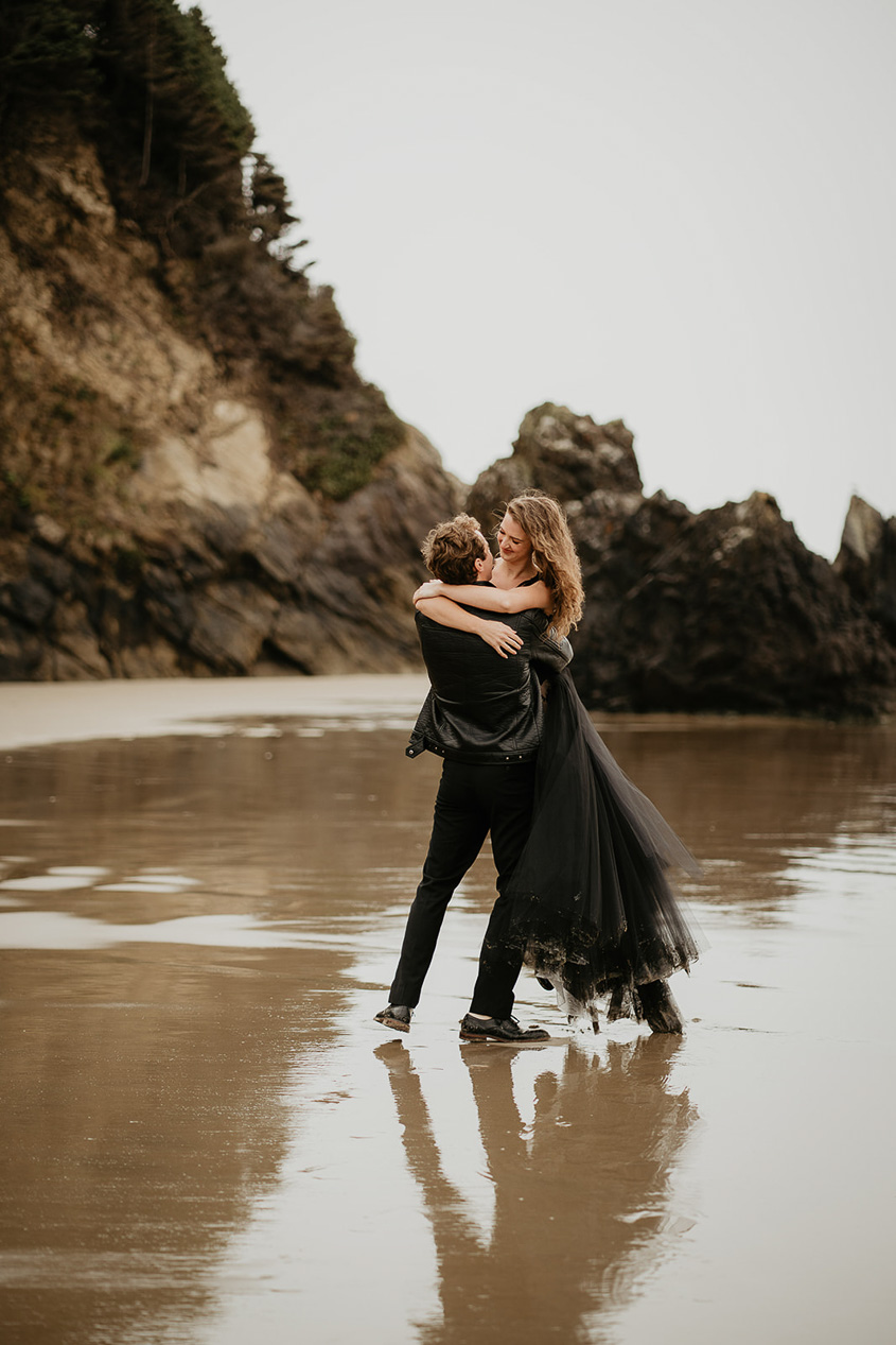 A couple wearing all black and hugging on the beach.