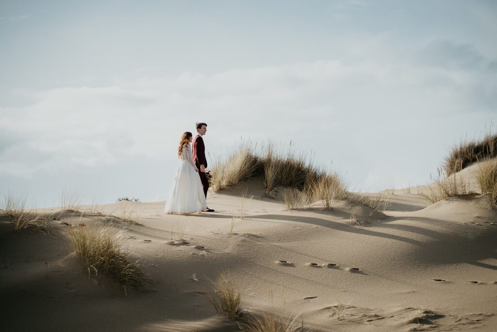 Bride and groom hold hands while walking across the sand dunes in Oregon