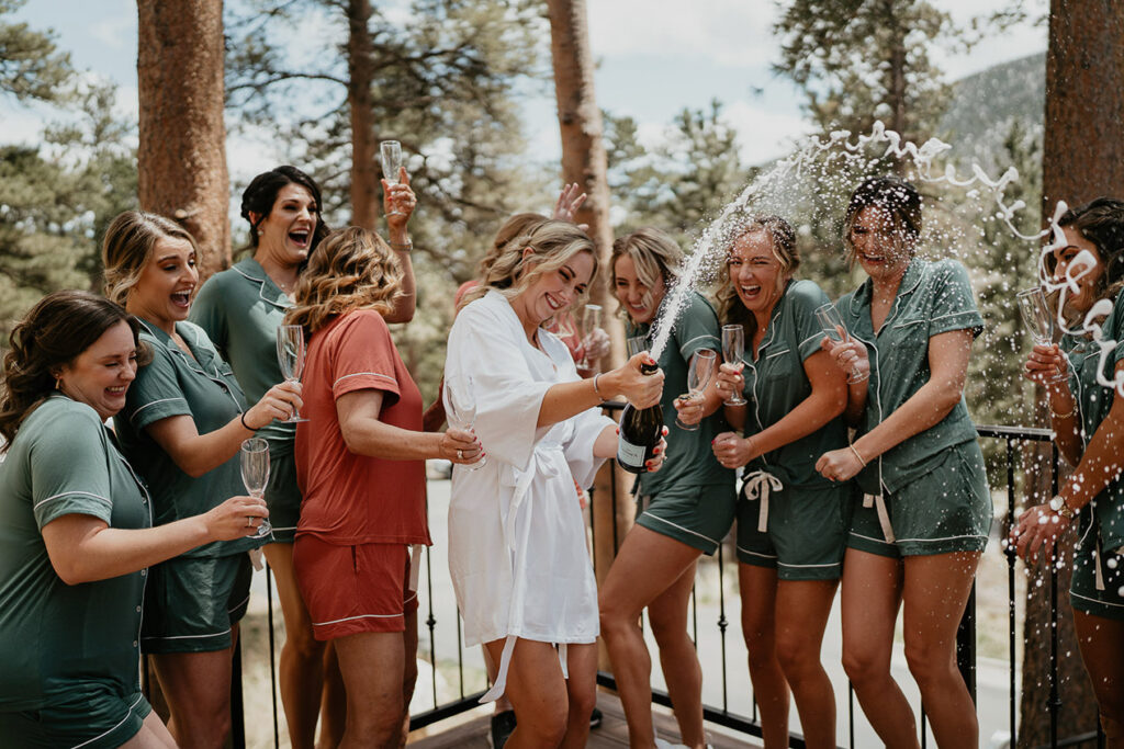 The bride opening up a bottle of champagne on a balcony with her bridesmaids and with pine trees in the background. 