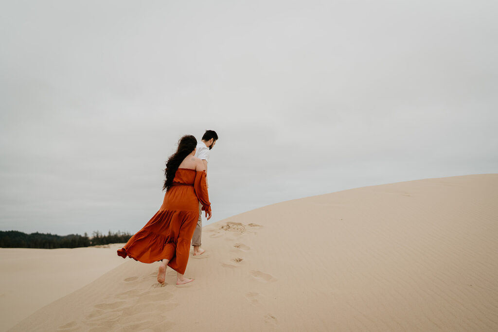 The fiance leading his partner across sand dunes in oregon. 