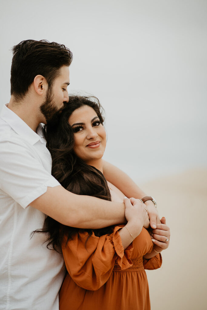 A boyfriend kissing his girlfriend on the head while she looks lovingly into the camera. 