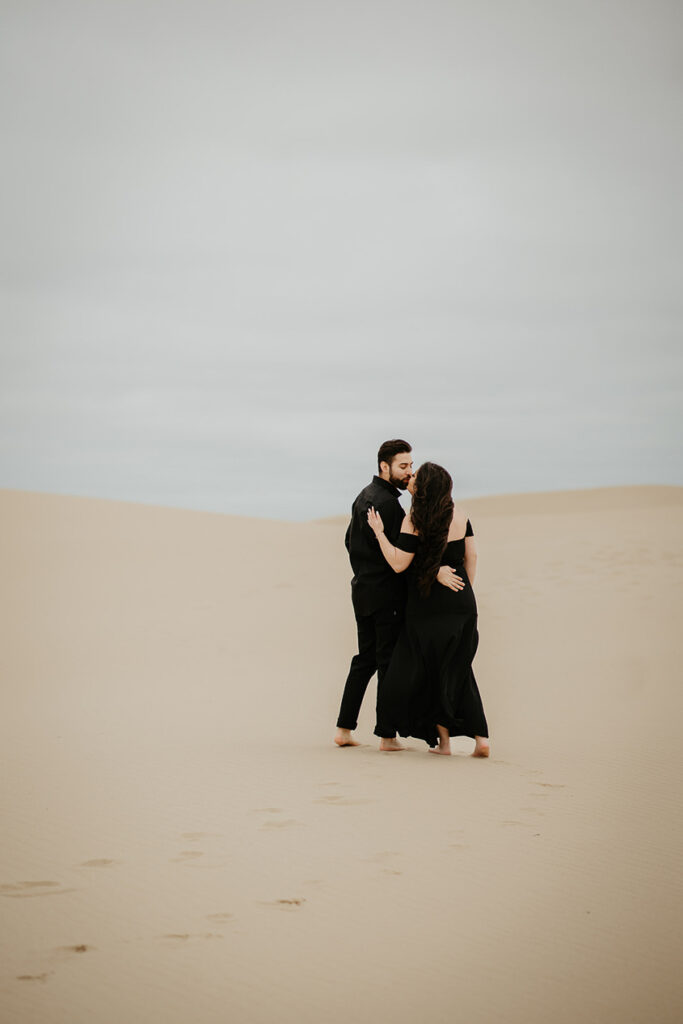 A couple kissing while walking barefoot on the Oregon Coast Sand Dunes and wearing al black. 