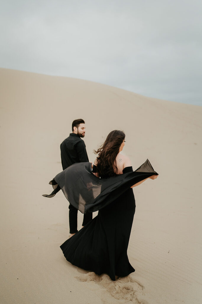 A couple, one with a black dress flowing in the wind and the other in a black button up and pants, walking along the Oregon Coast Sand Dunes during their engagement session. 