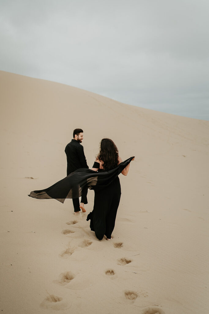 A couple, one with a black dress flowing in the wind and the other in a black button up and pants, walking along the Oregon Coast Sand Dunes during their engagement session. 