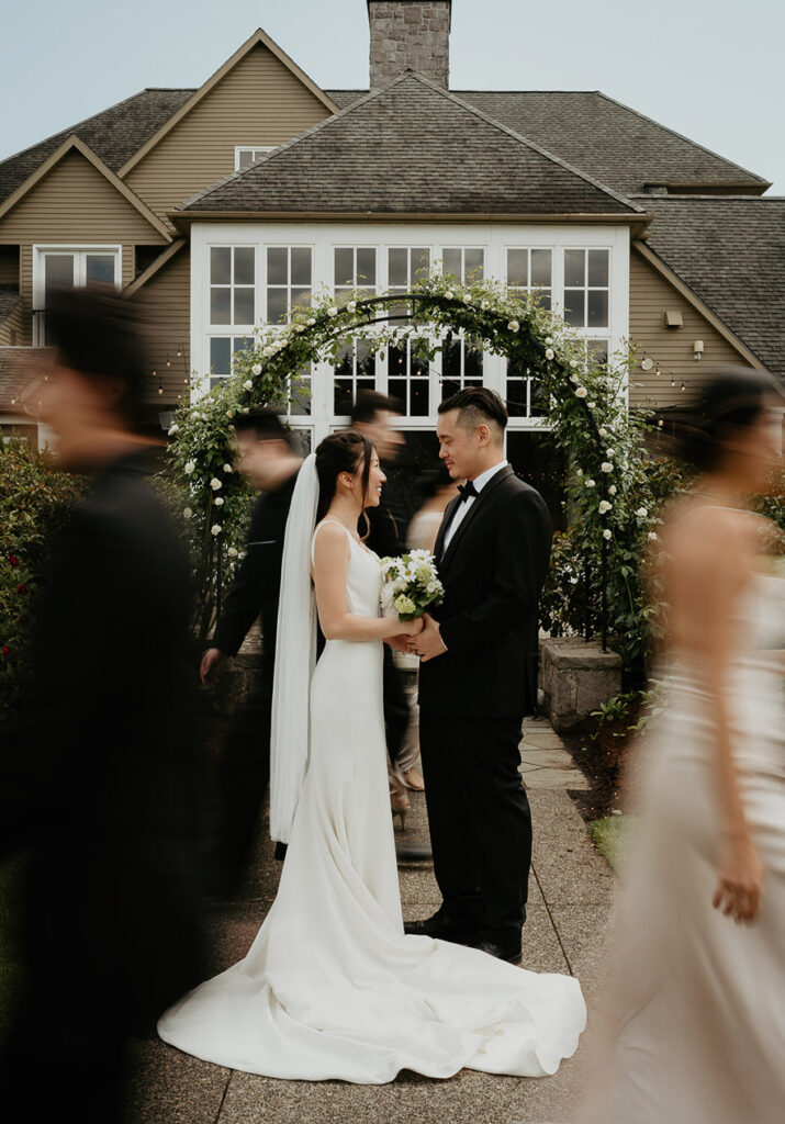 Judith and Joshua holding each other under a flowery, green arch with the blurry wedding party walking past. 