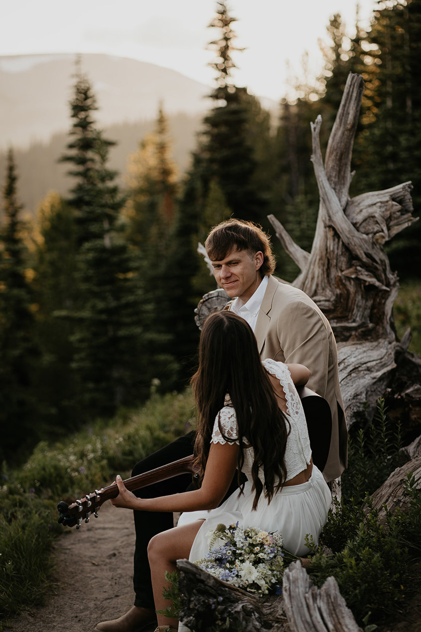 The wife playing guitar for her husband sitting on a fallen tree. 