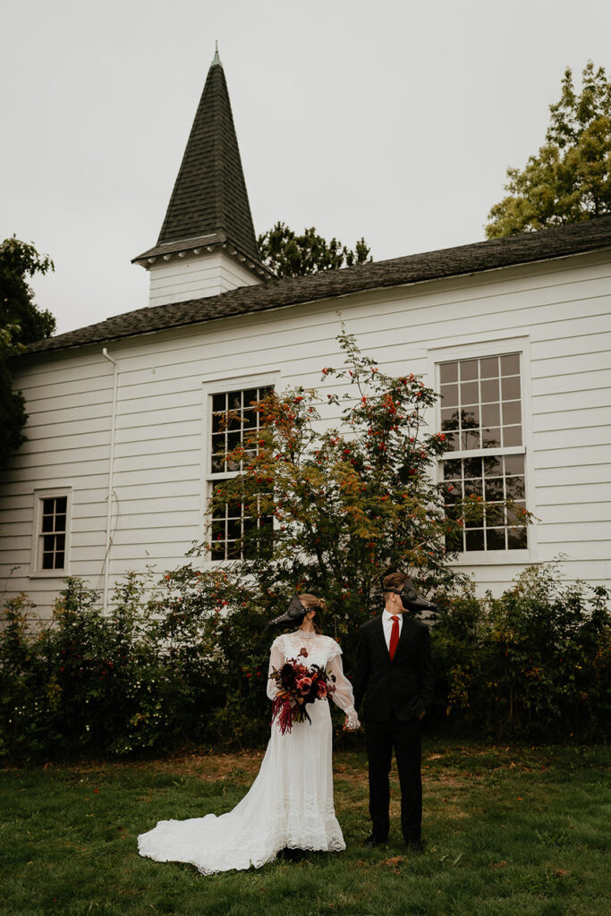 A couple wearing bird masks holding hands in front of a chapel. 