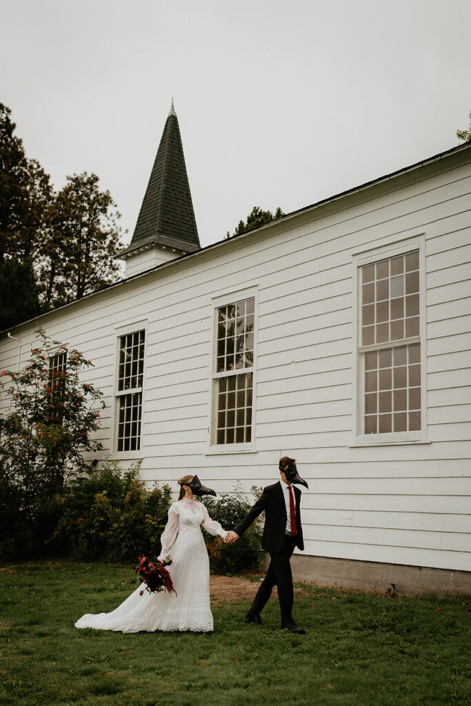 A couple wearing bird masks in front of a chapel, the groom leading the wife. 