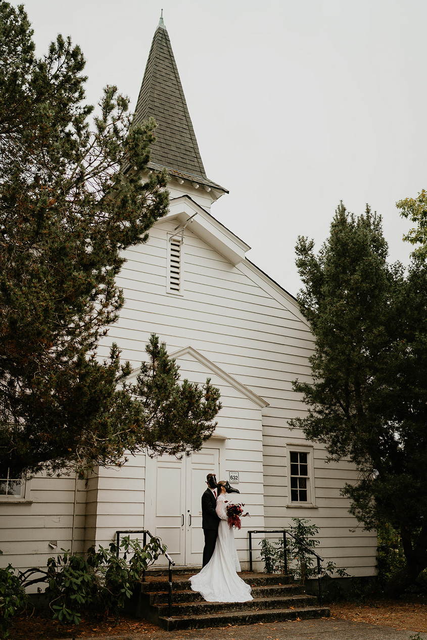 A couple wearing bird masks holding each other in front of a chapel. 