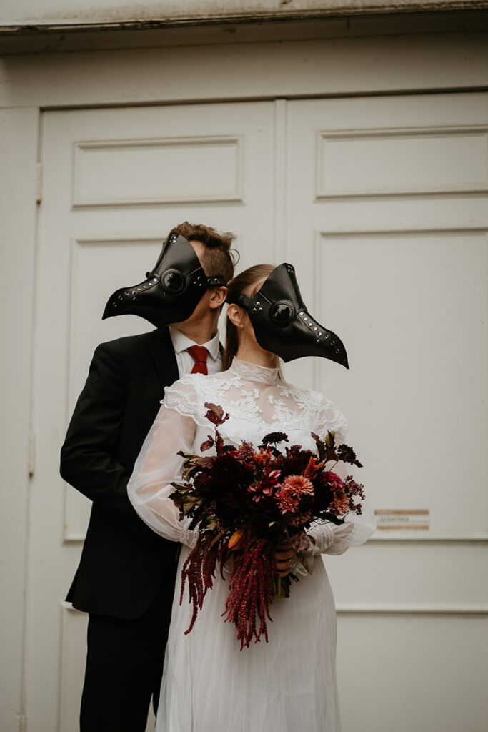 A couple wearing bird masks holding each other in front of a chapel. 