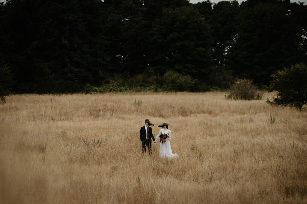 A couple wearing bird masks holding hands standing in an open field. 