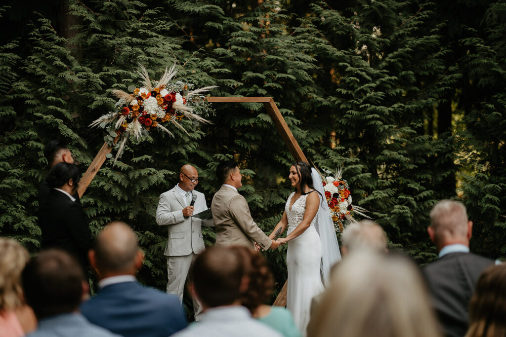 The brides holding hands as the officiant makes a speech, with wedding guests onlooking.