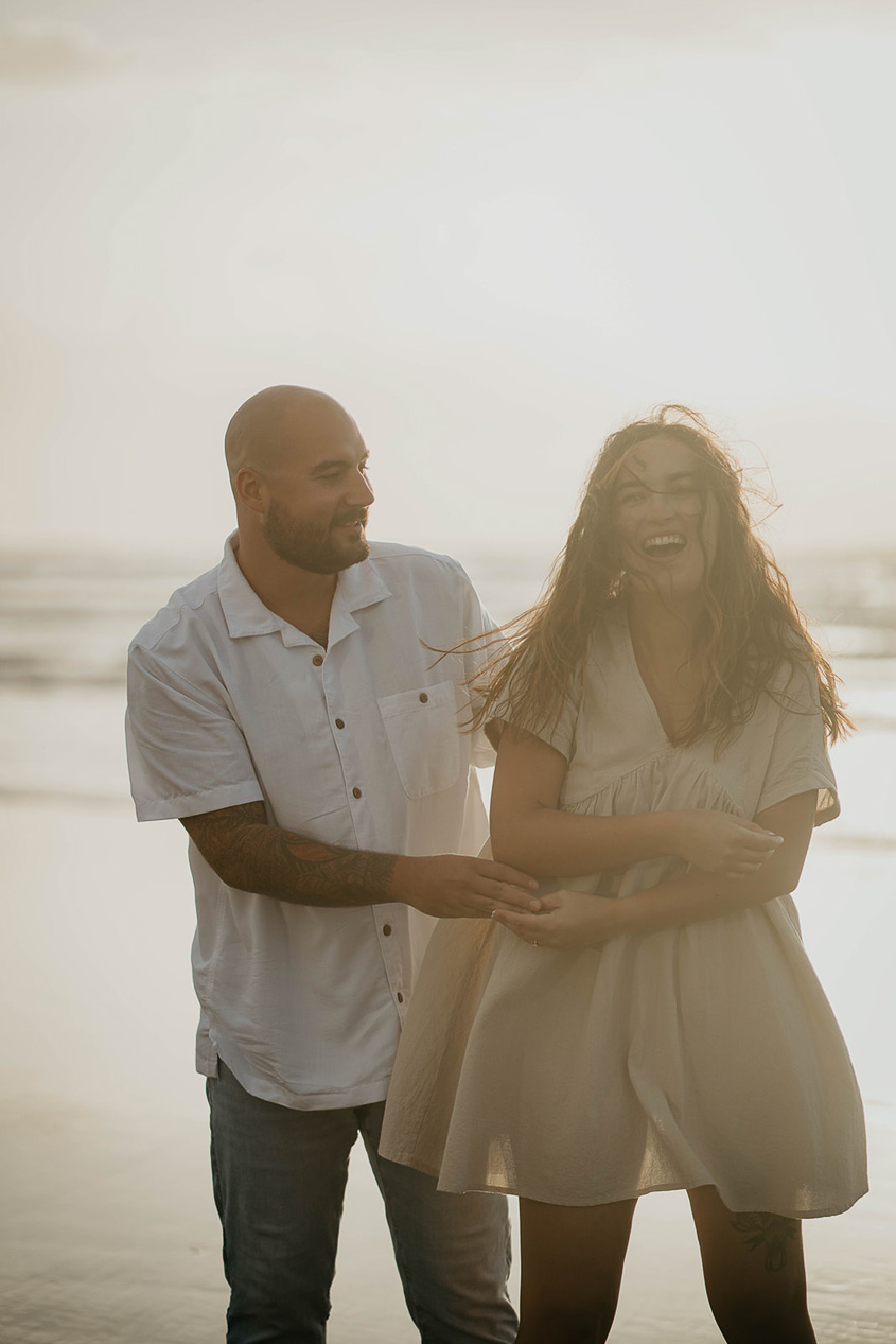 a newly engaged couple laughing with the Pacific Ocean in the background. 