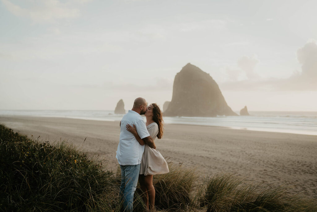 a newly engaged couple kissing among beach grass with Haystack Rock in the background. 