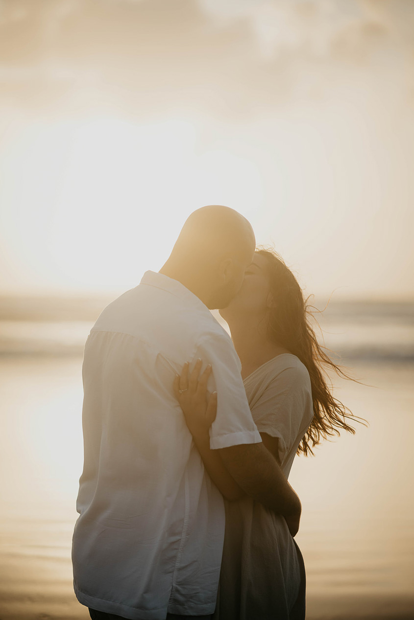 a couple kissing on the beach with the sun setting in the background.