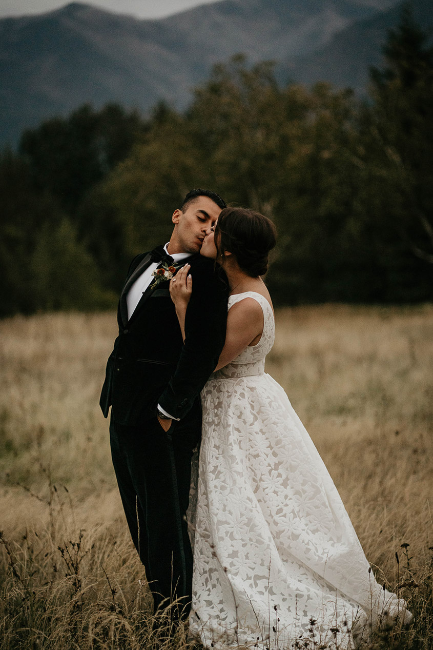 the newlyweds kissing with a field and pine trees in the background. 