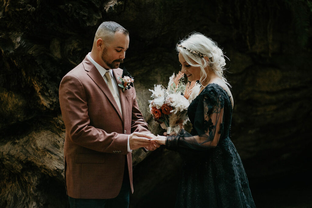 The husband putting on her wife's ring in front of a redwood tree. 