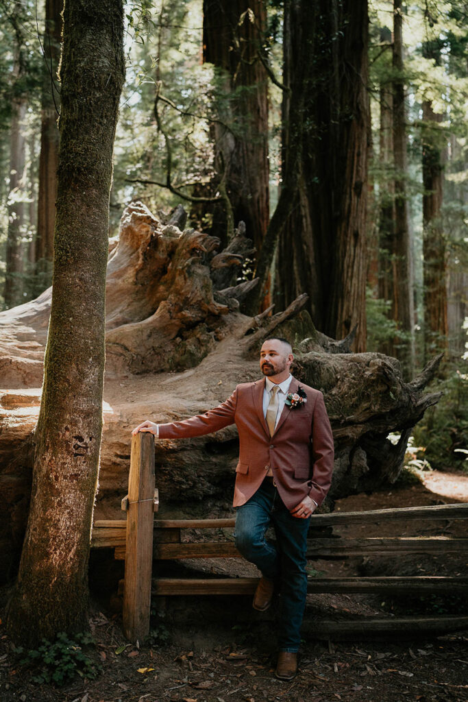 The husband standing against a fence with redwood trees behind him. 