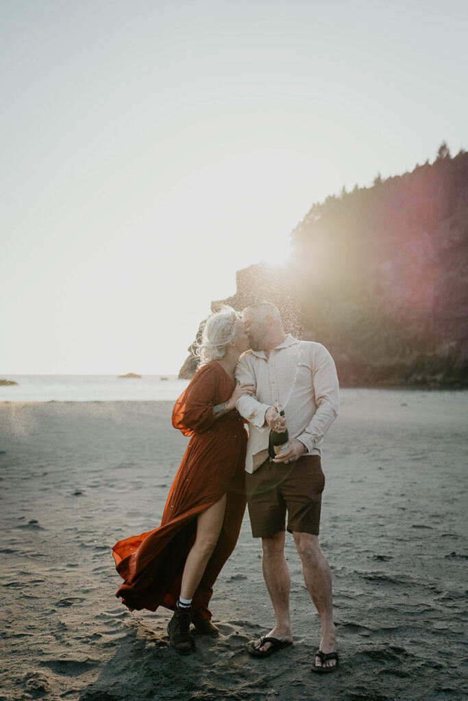 the husband and wife kissing on the beach while popping a bottle of champagne. 