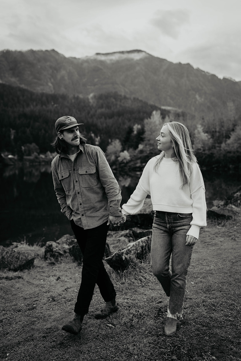 A couple holding hands and looking lovingly at each other with the Columbia river and evergreen trees in the background. 