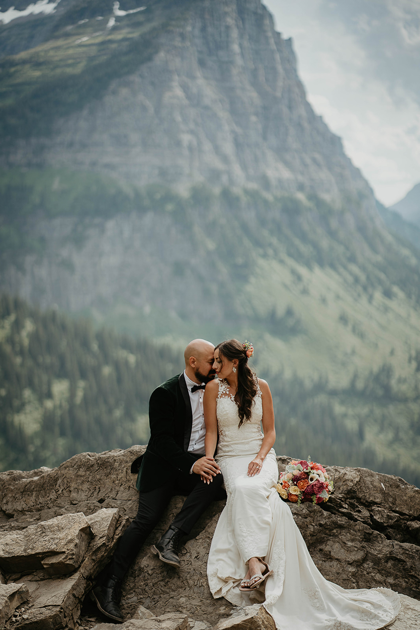 the newlyweds sitting on a rock and embracing each other with large mountains surrounding them. 