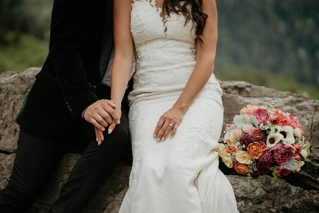 The couple sitting down and holding hands with a bouquet of flowers next to the bride. 